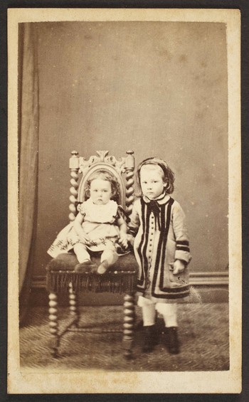 Unknown sitter [portrait of seated infant on upholstered chair left to a standing girl]